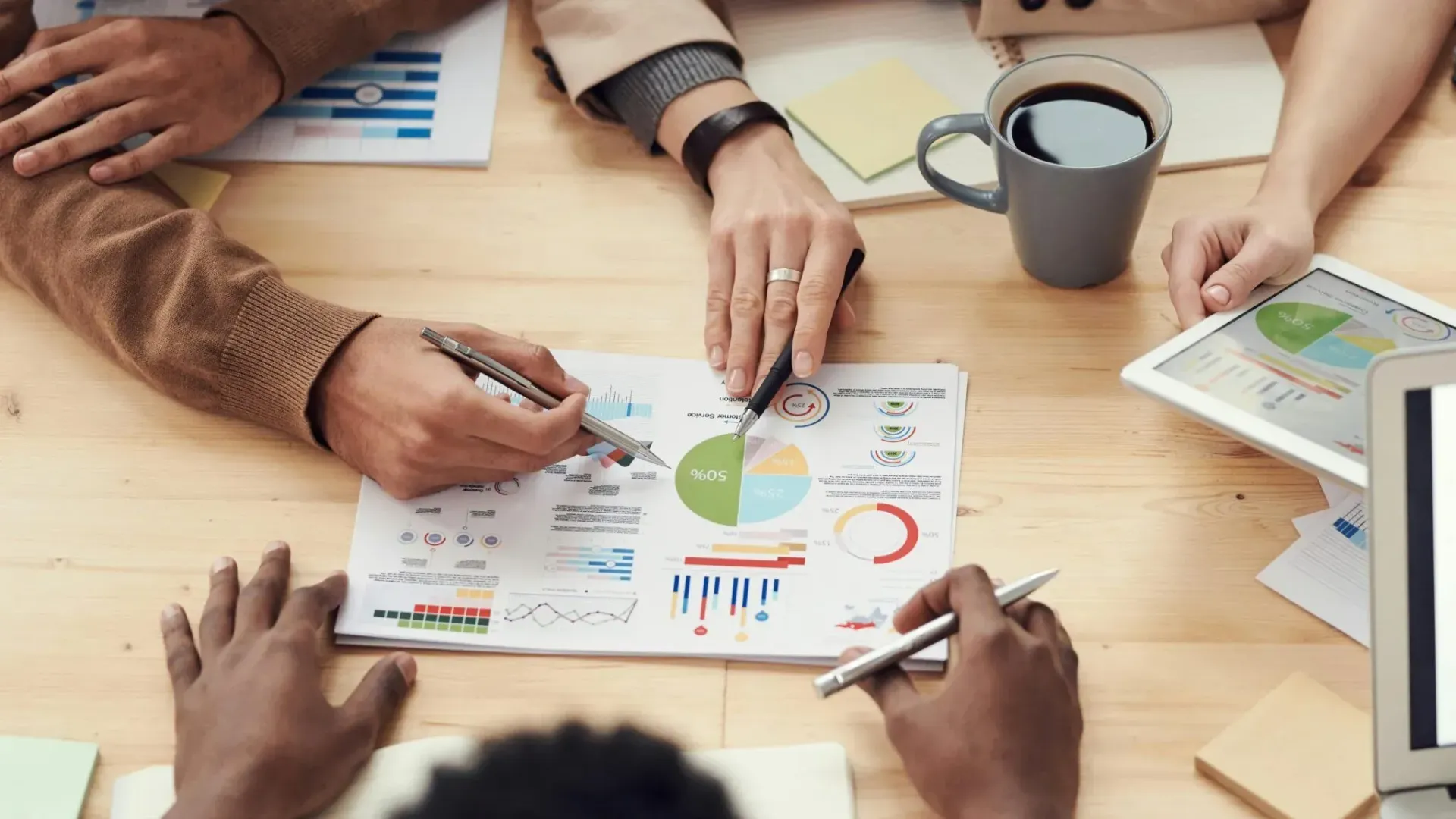Hands around a table reviewing charts and pie graphs with a coffee mug nearby.