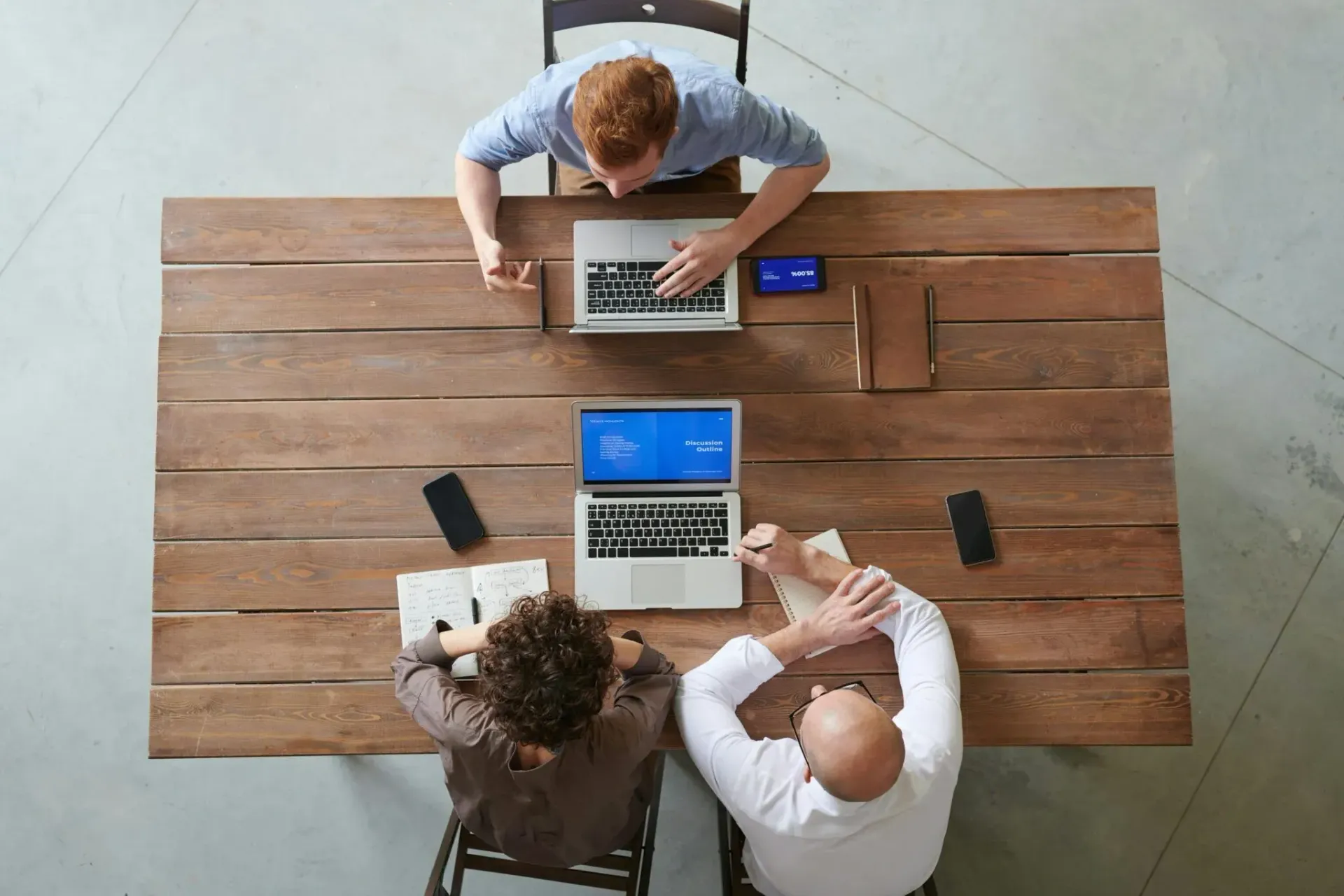 Three people working on laptops around a wooden table in a meeting room, overhead view