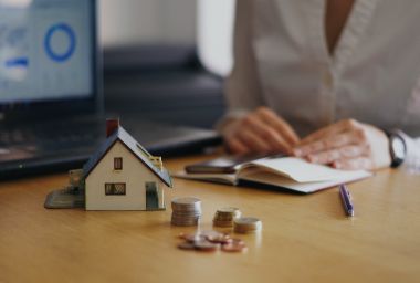 Miniature house, coins, and notebook on a desk with a person in the background, possibly doing financial planning.
