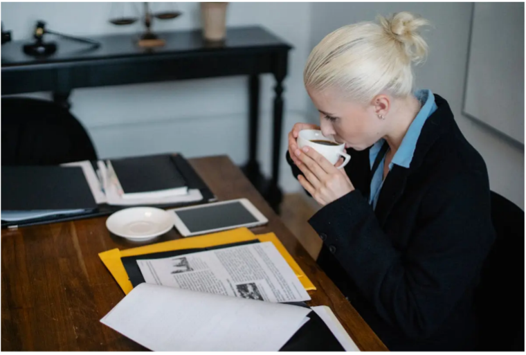Woman in blazer drinks coffee at a desk with documents and a tablet in a law office.