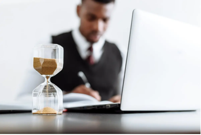 Hourglass on desk, man in vest working on laptop and writing.