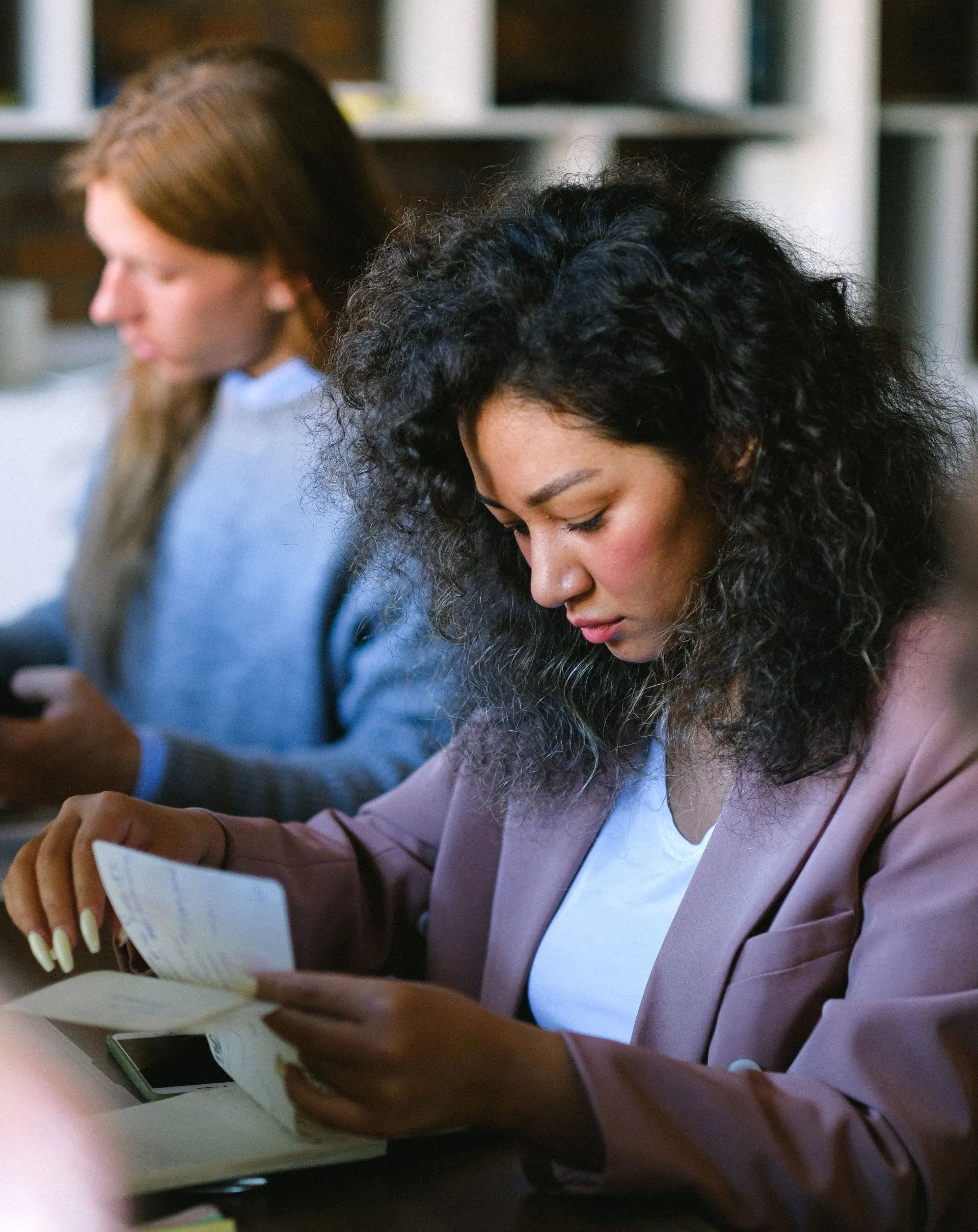 Woman with curly hair, in pink blazer, looking down at documents, another person blurred in background.