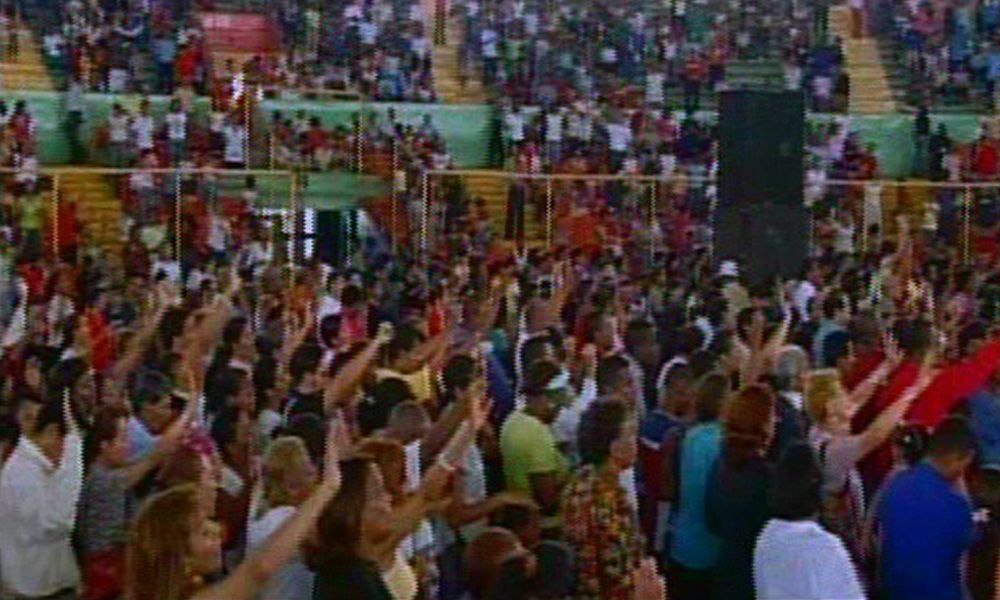 Large crowd in stadium with raised arms, likely worshiping. Many people, bright colors.