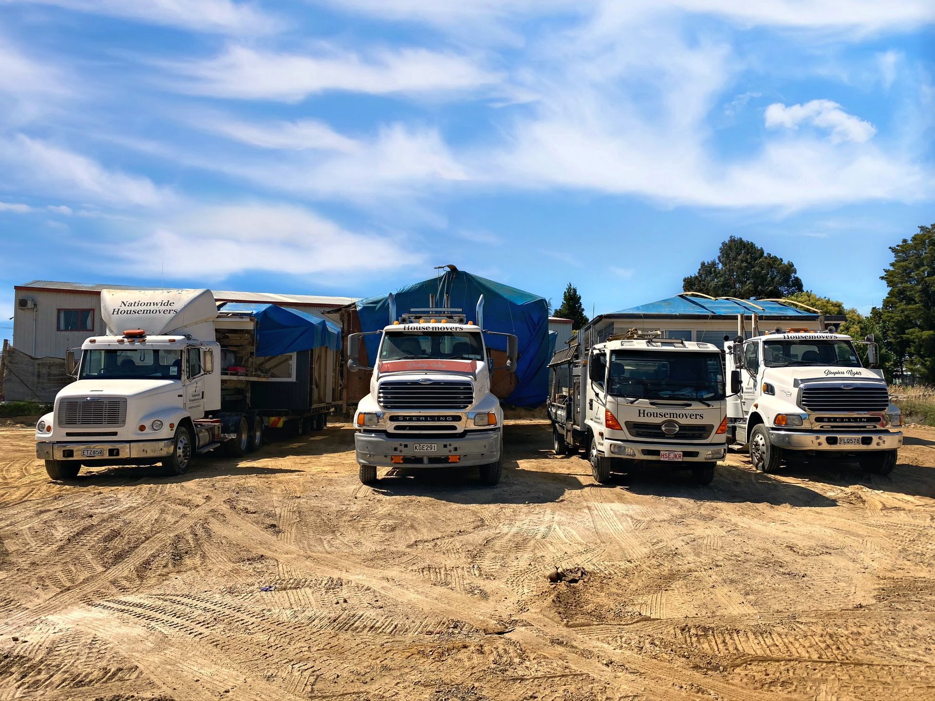 Five white dump trucks parked on a dirt lot under a blue sky with scattered clouds