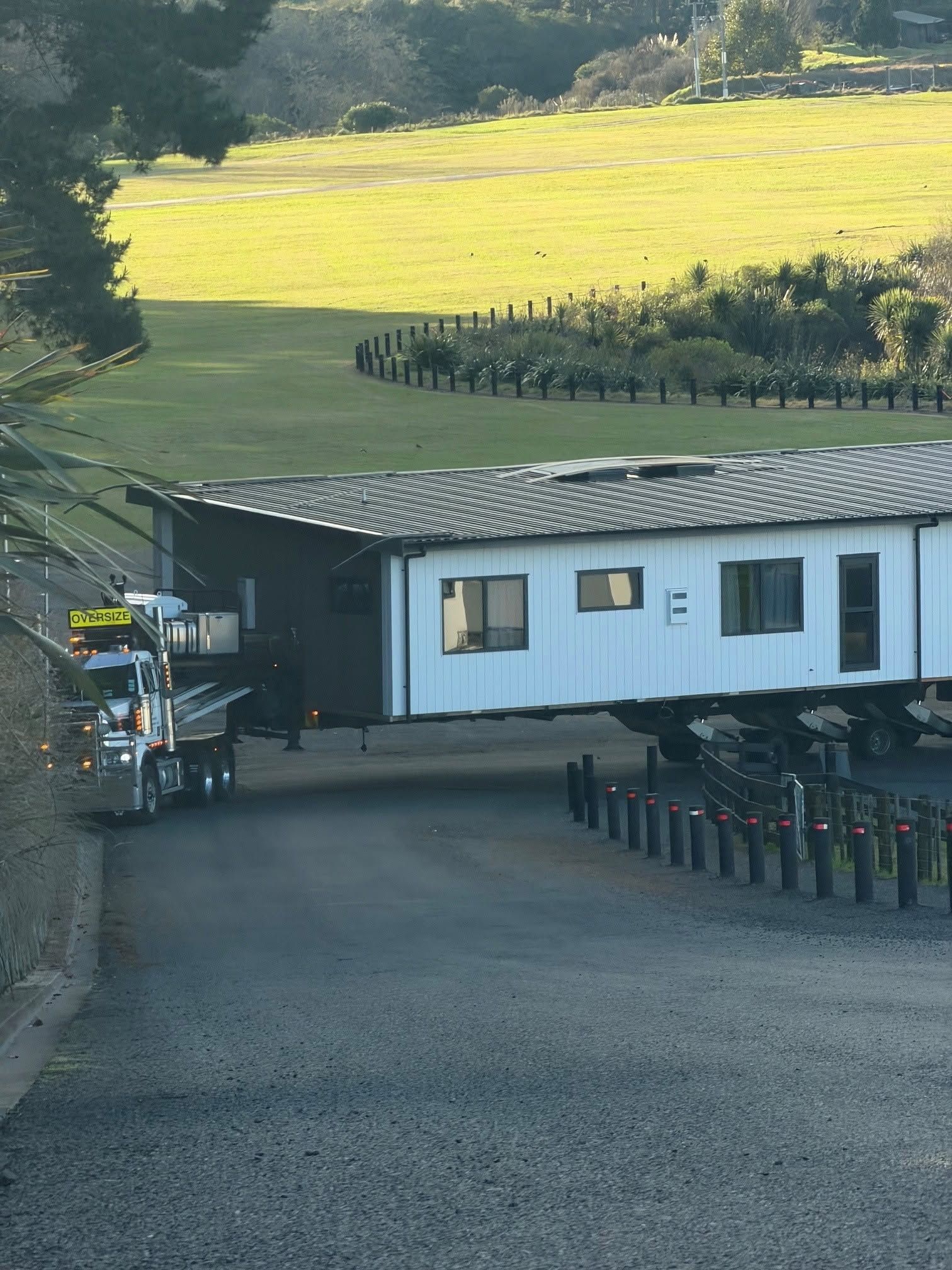 White building with a dark roof beside a gravel driveway, with sunlit fields and trees in the background