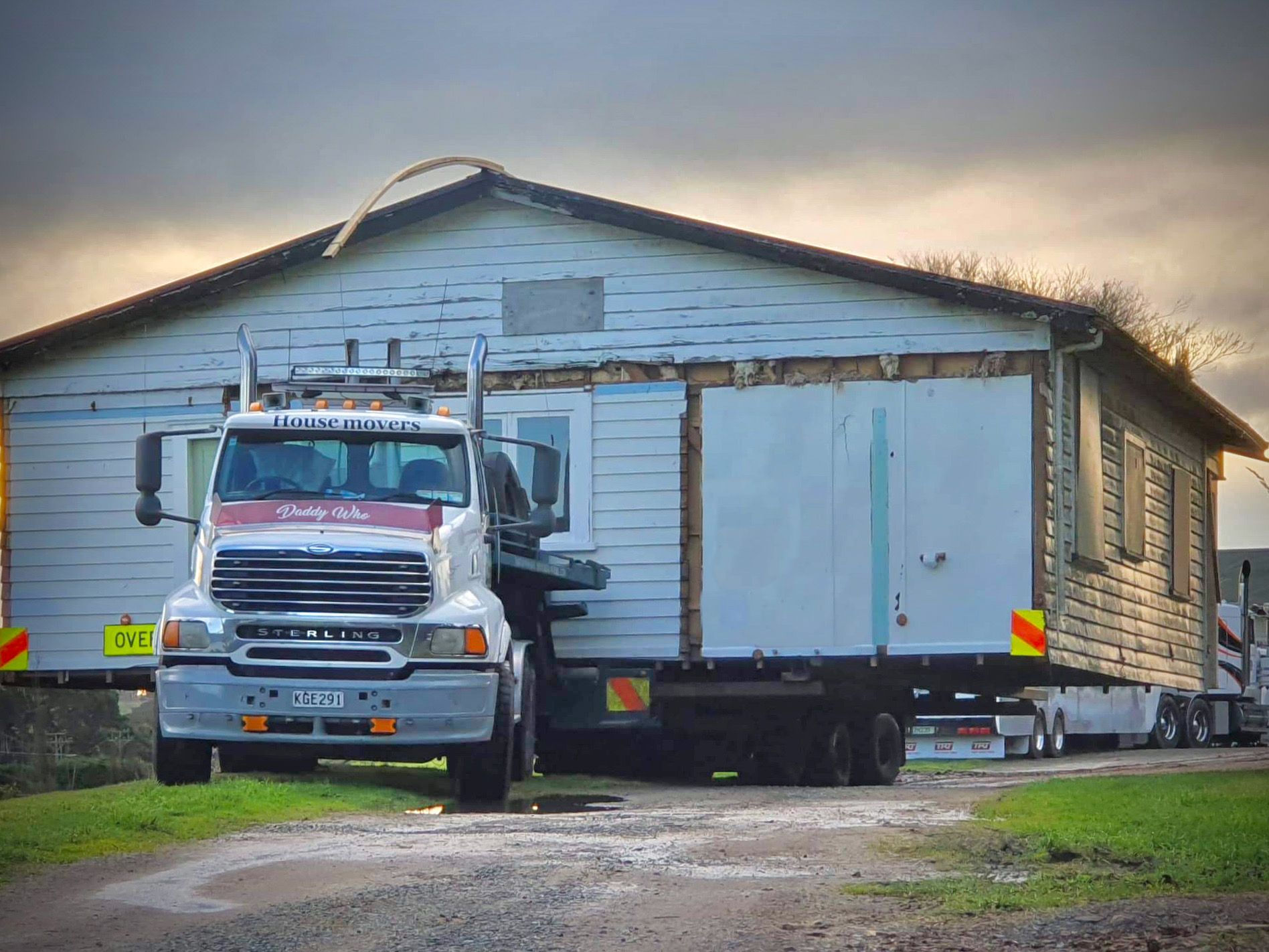 House being moved on a truck along a road, with a white trailer attached to the structure.