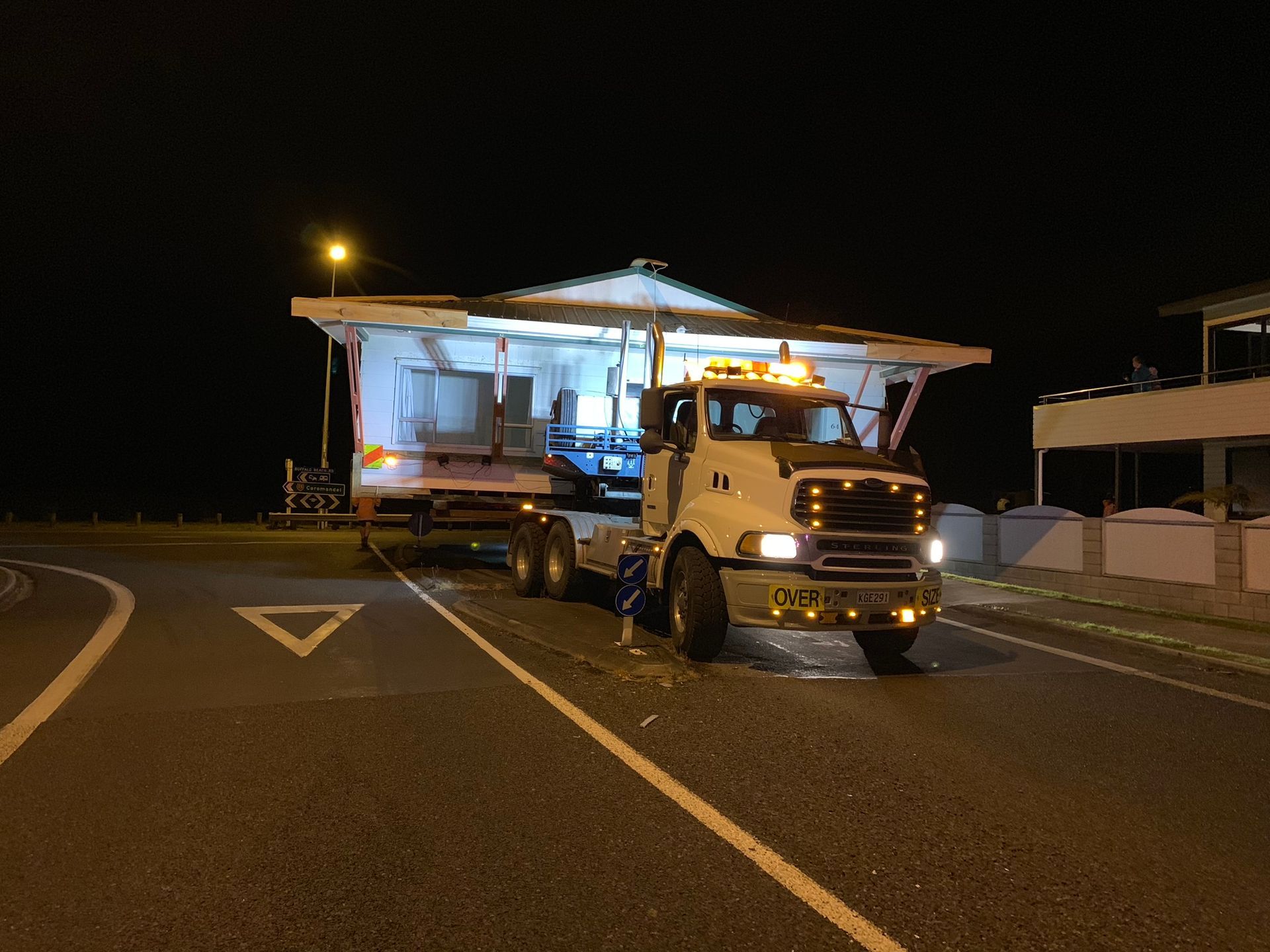 Night road scene with a lit flatbed truck carrying a small house-like structure.
