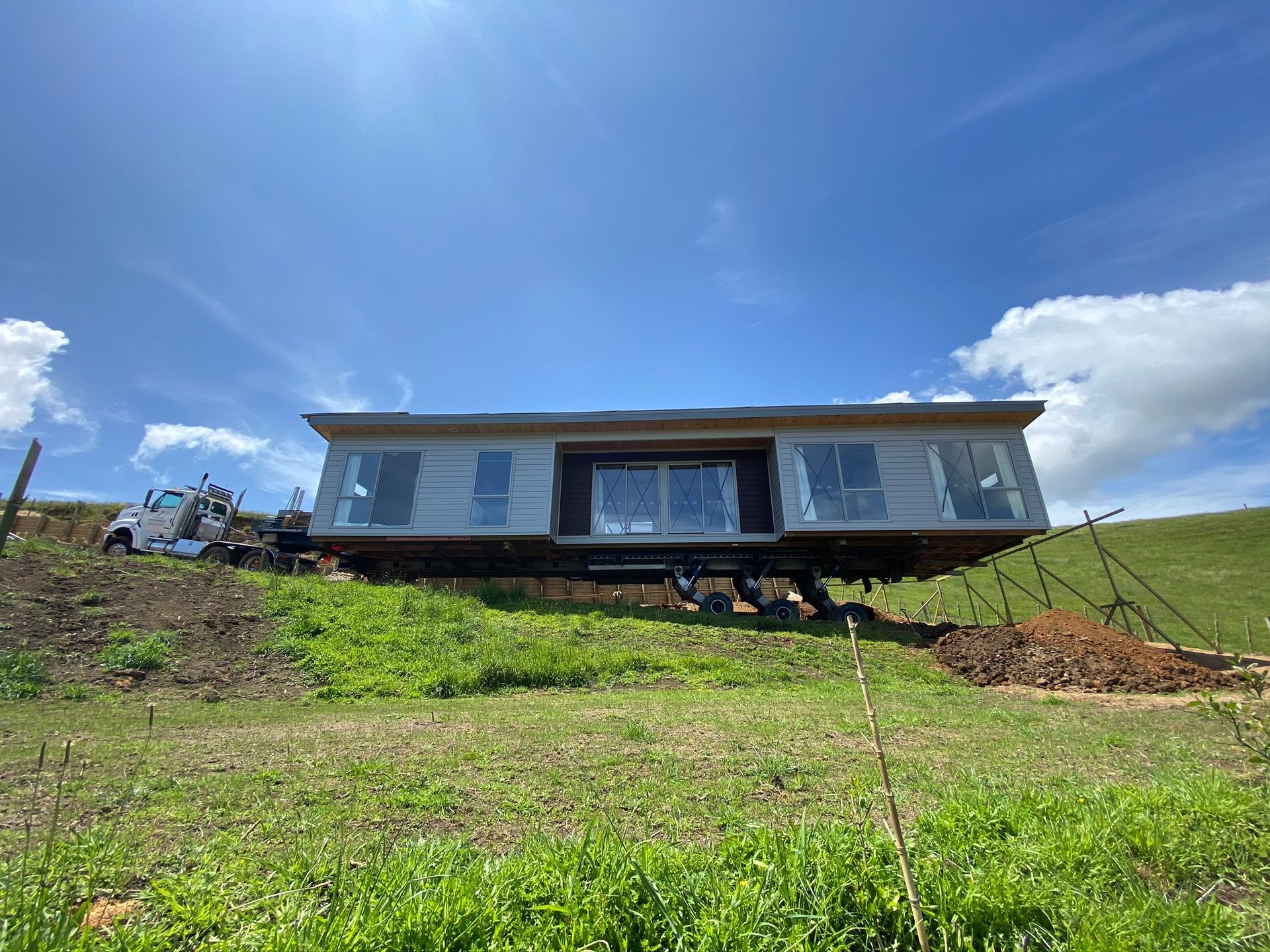 Single-story house with a metal roof on a grassy hillside under a blue sky with clouds