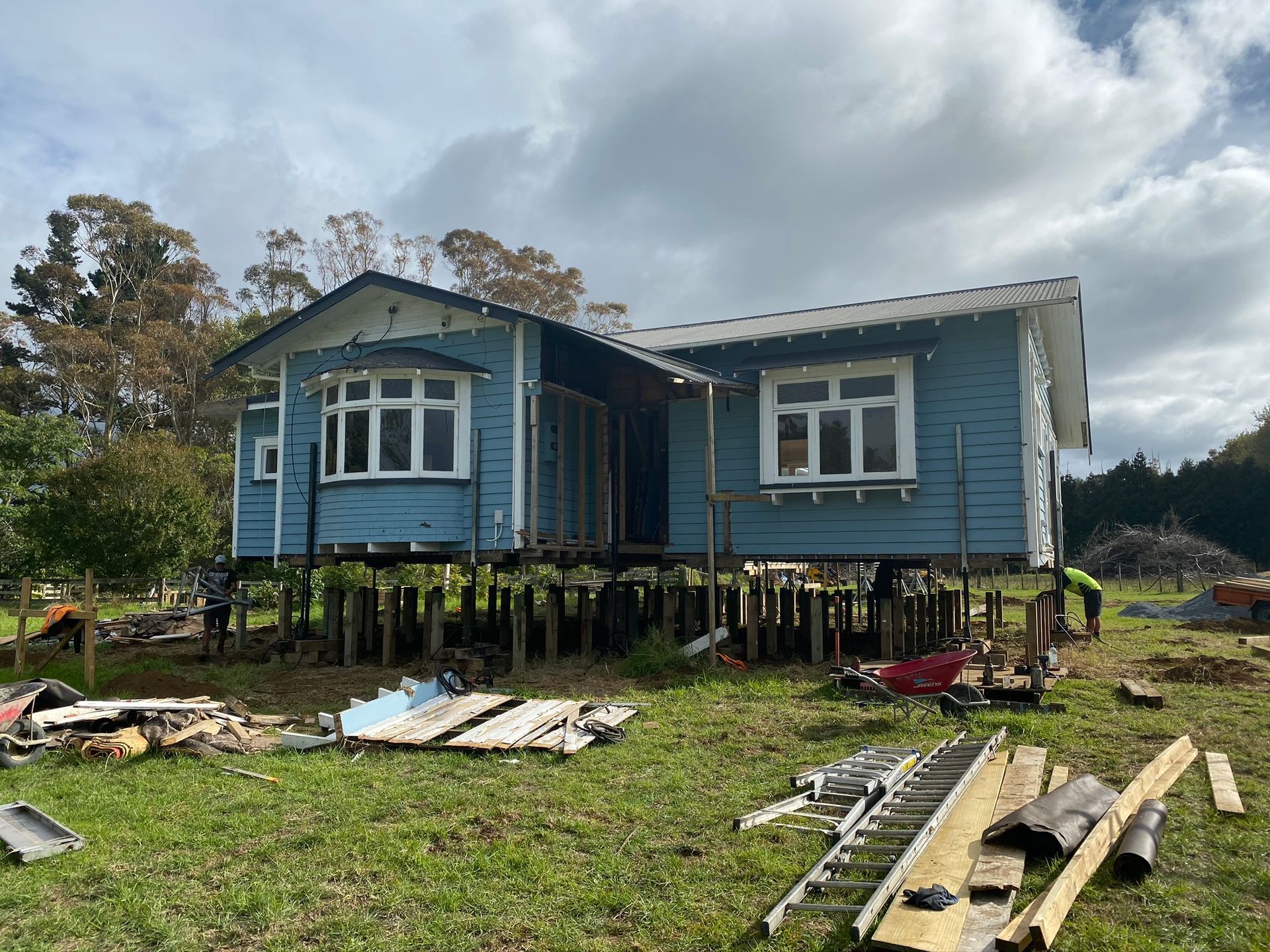 Blue stilt house under repair, raised on posts in a grassy yard with construction materials scattered nearby.