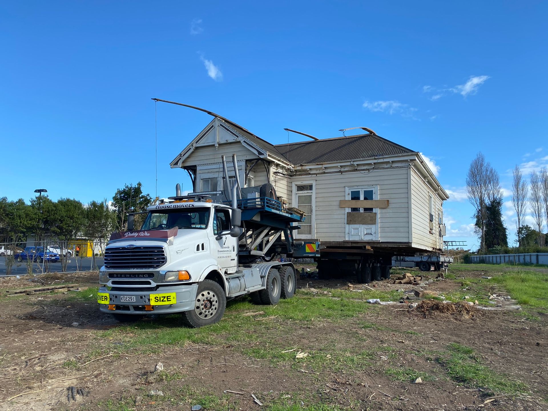 Tow truck lifting a weathered white house on a flatbed in a rural field under a blue sky
