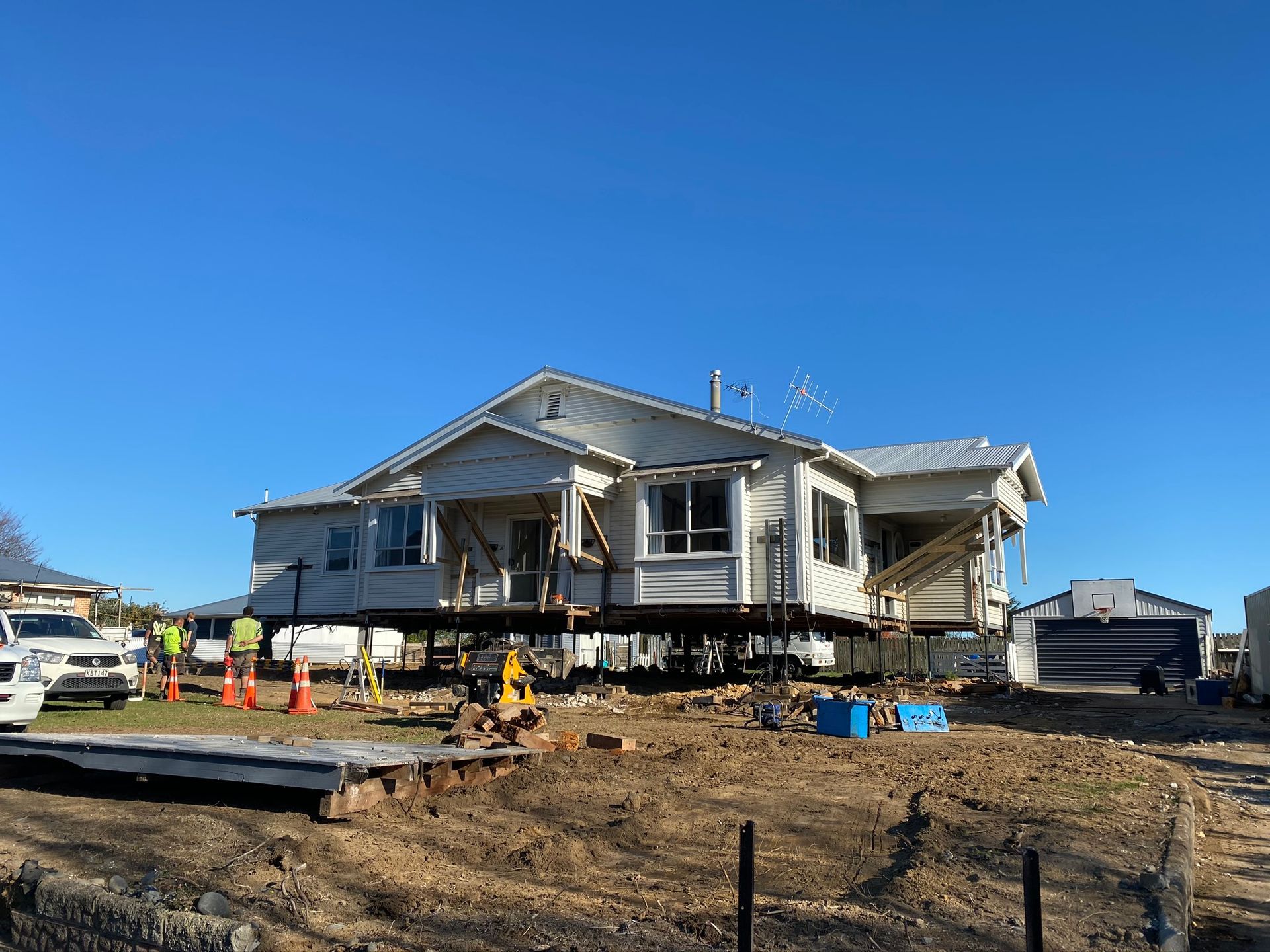 House lifted on construction jacks at a worksite, with workers and equipment under a clear blue sky