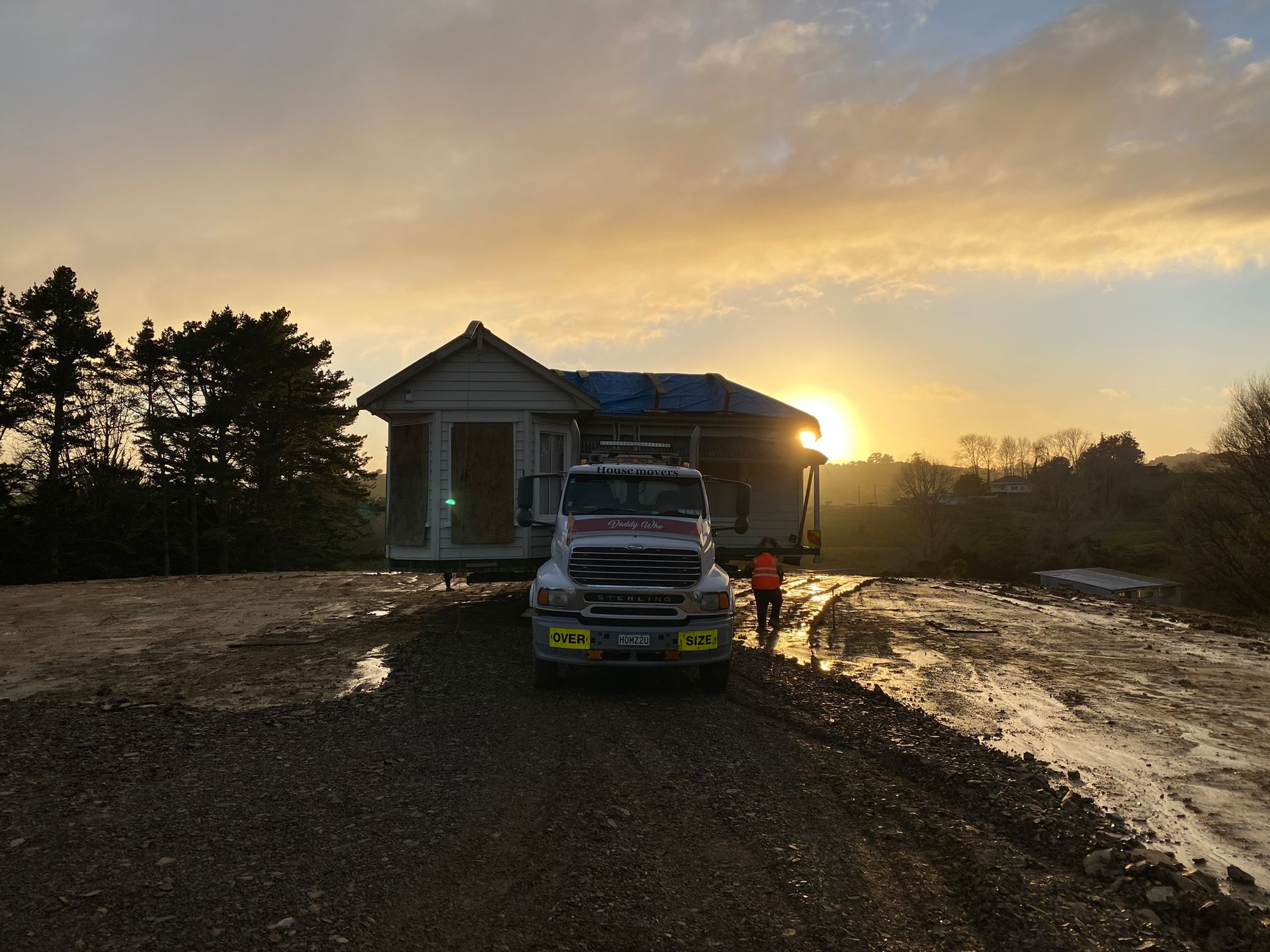 Moving truck loaded with belongings parked in front of a house at sunset, with a person standing nearby.