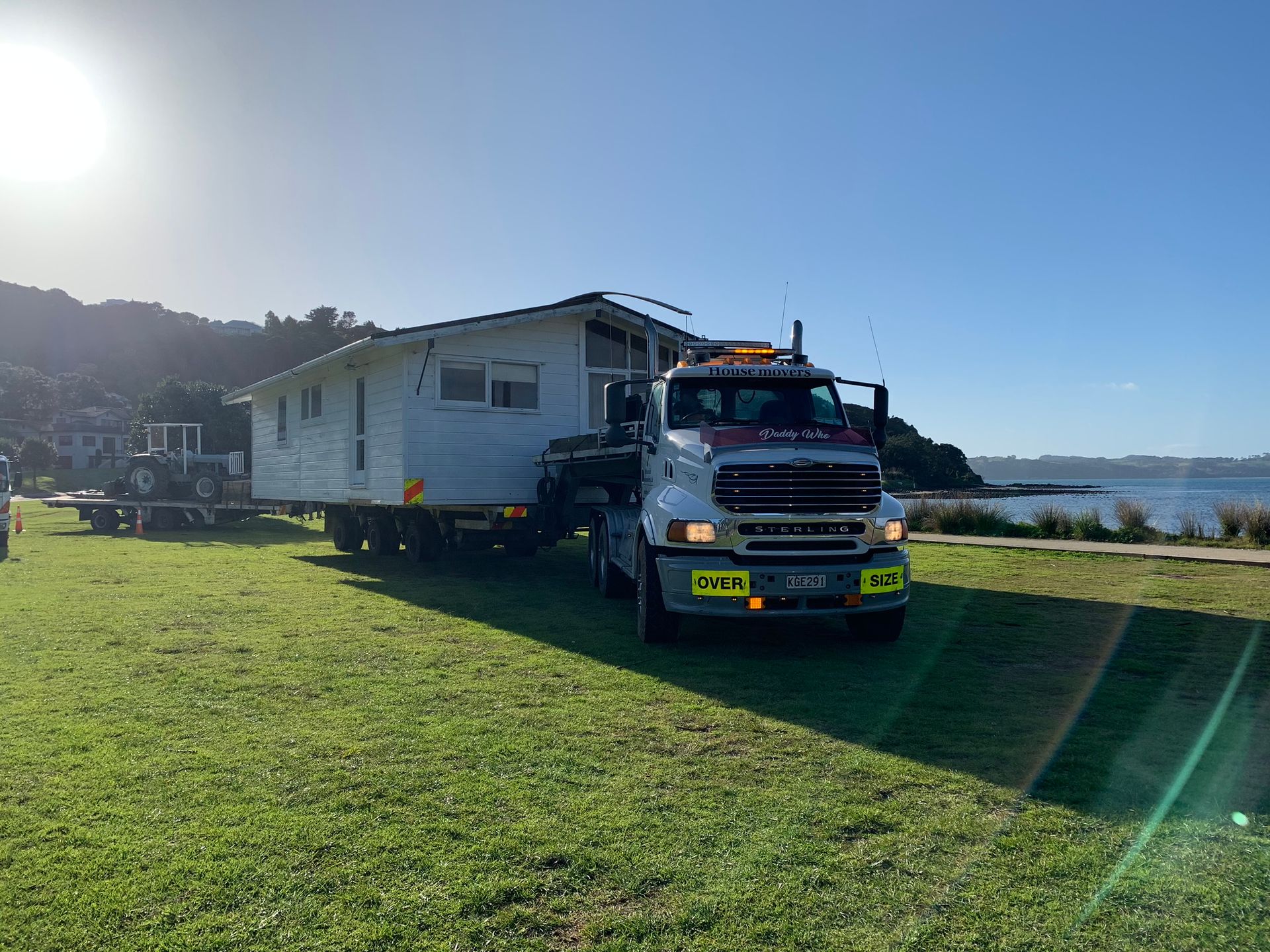 Tow truck hauling a white mobile home across a sunny grassy field near the water