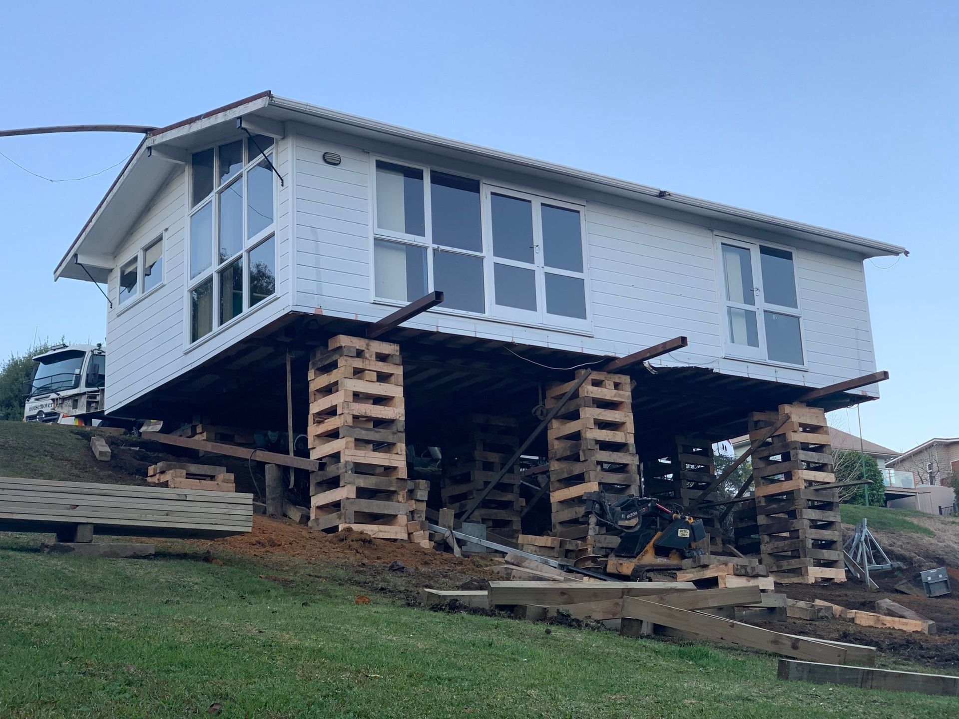 House elevated on stacked timber supports over a sloped yard, with damaged debris below.