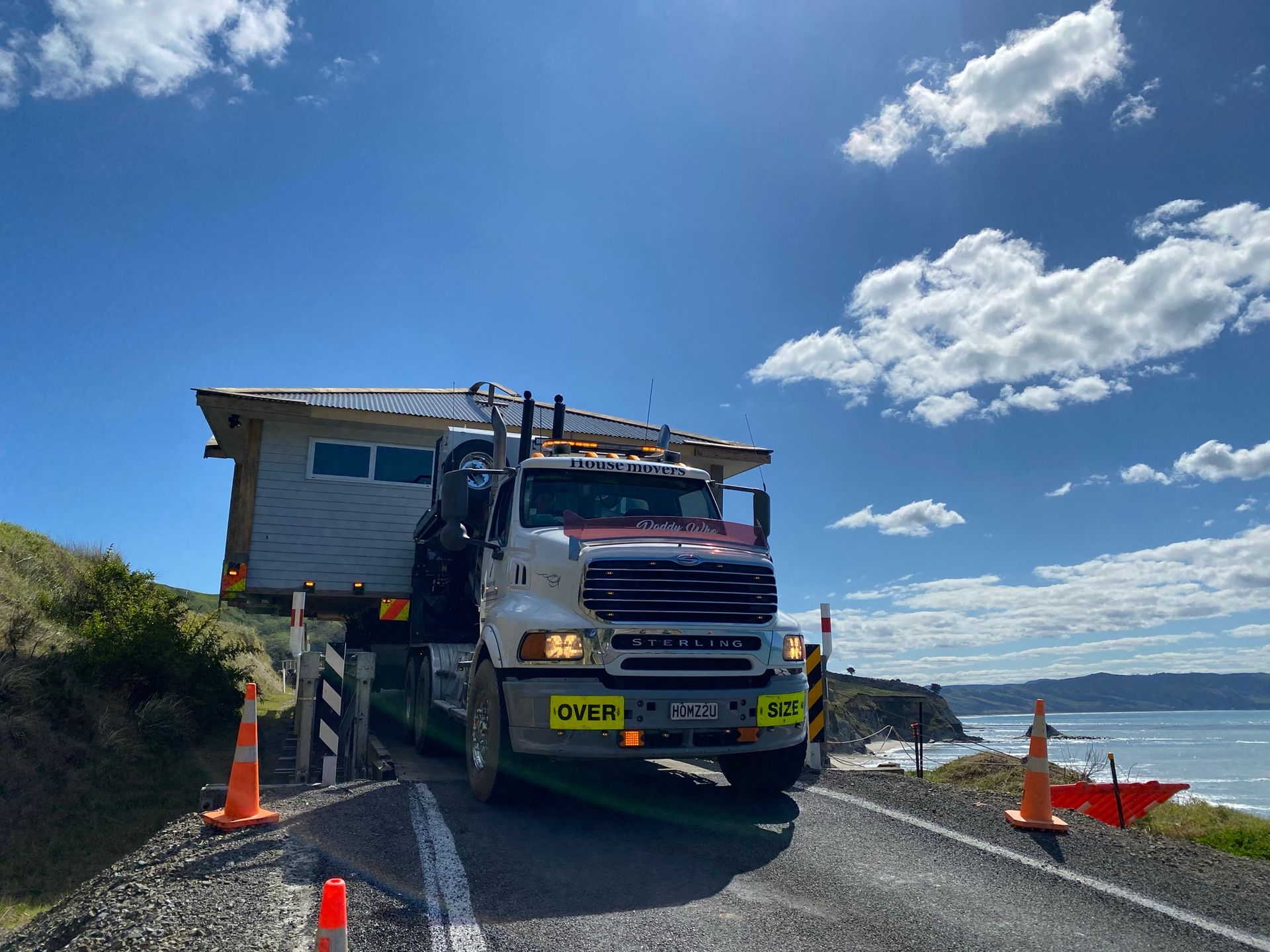 Tow truck towing a house on a rural road under a blue sky with clouds