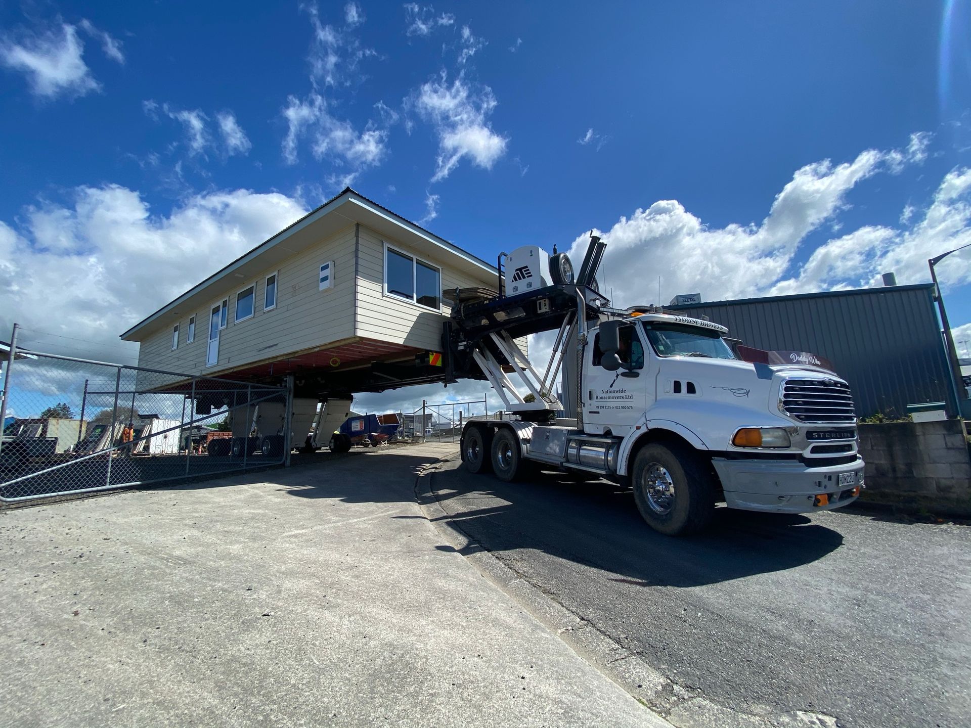 Tow truck lifting a beige house module in a gravel lot under a blue sky