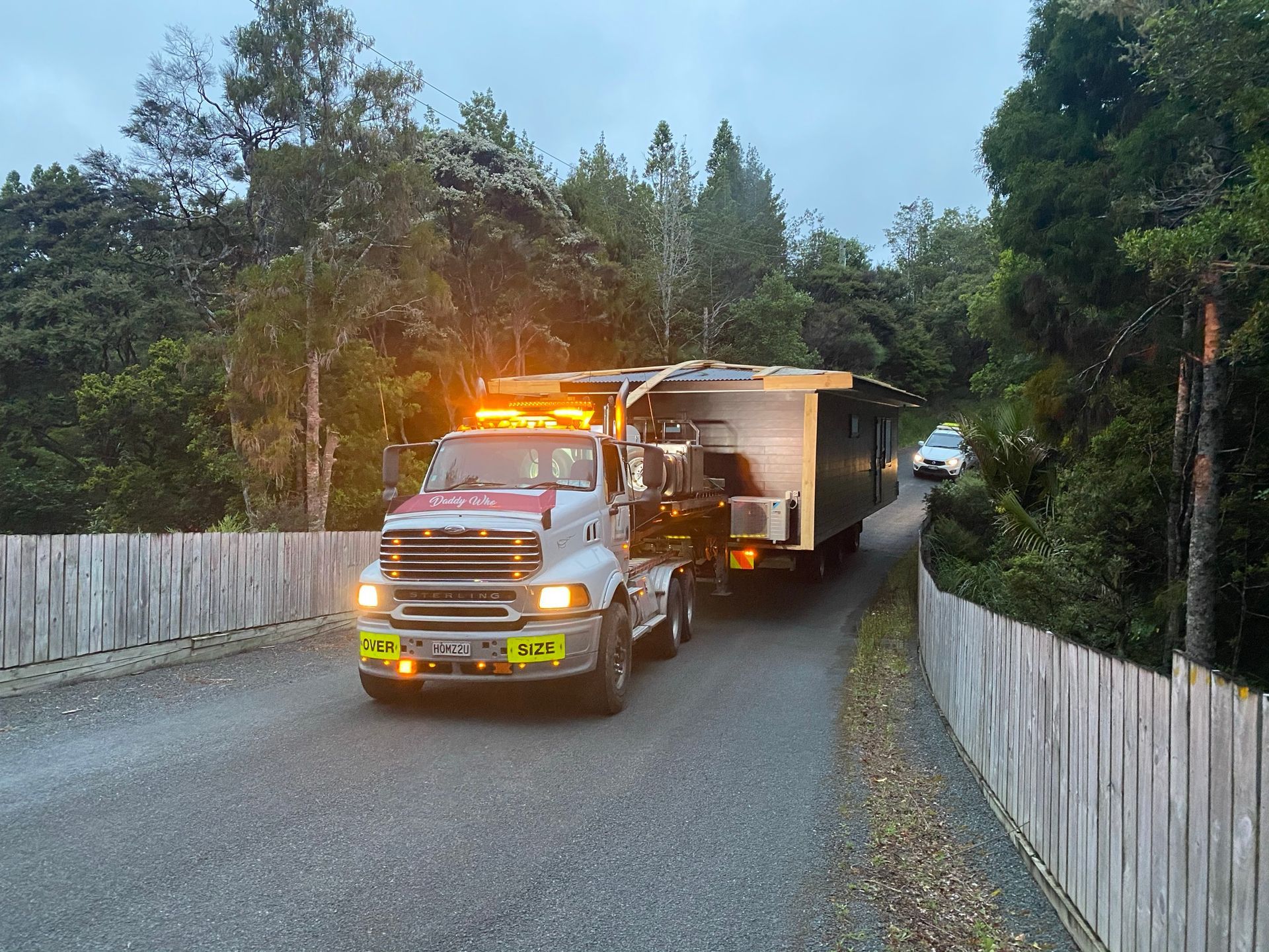 Tow truck towing a trailer on a narrow rural road at dusk, surrounded by trees and fences