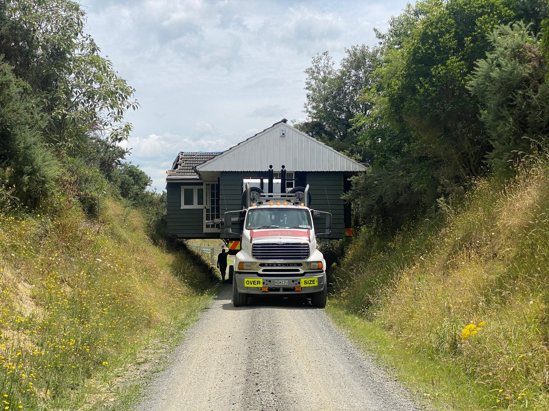 Truck driving down a narrow gravel lane toward a dark house, flanked by tall grass and trees