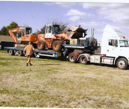 Orange construction loader on a flatbed truck, with a worker nearby in a grassy field.