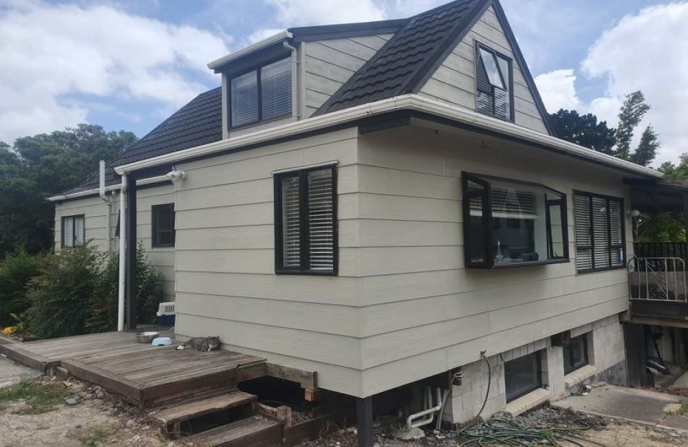 Cream house with dark roof and dormer windows, viewed from the side in a residential yard.