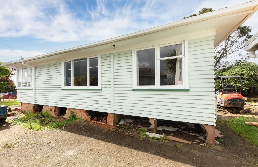 Single-story house on stilts with pale green siding and white trim, shown in a yard.