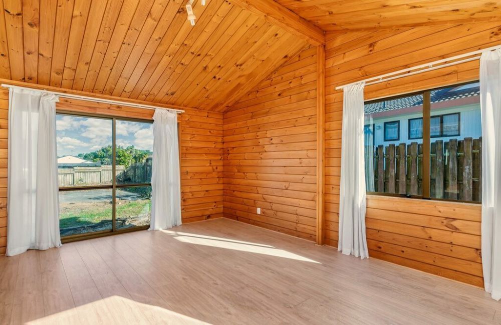 Empty wood-paneled room with large windows, white curtains, and a sunlit view outside