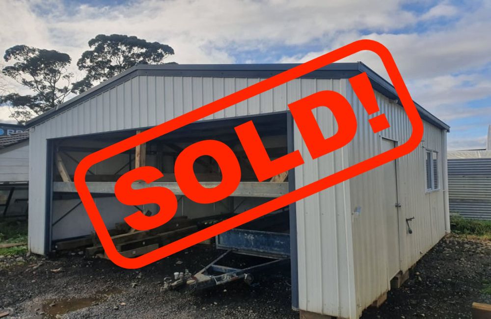 White shed with red SOLD sign in front, shown outdoors on gravel near trees