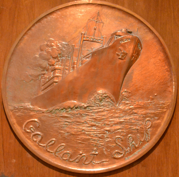 Bronze circular relief of a ship at sea, waves crashing around the hull. Text reads 