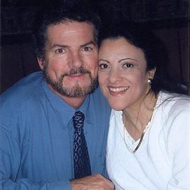 Man with beard and woman smiling, close together, indoors. Man in blue shirt and tie, woman in white.