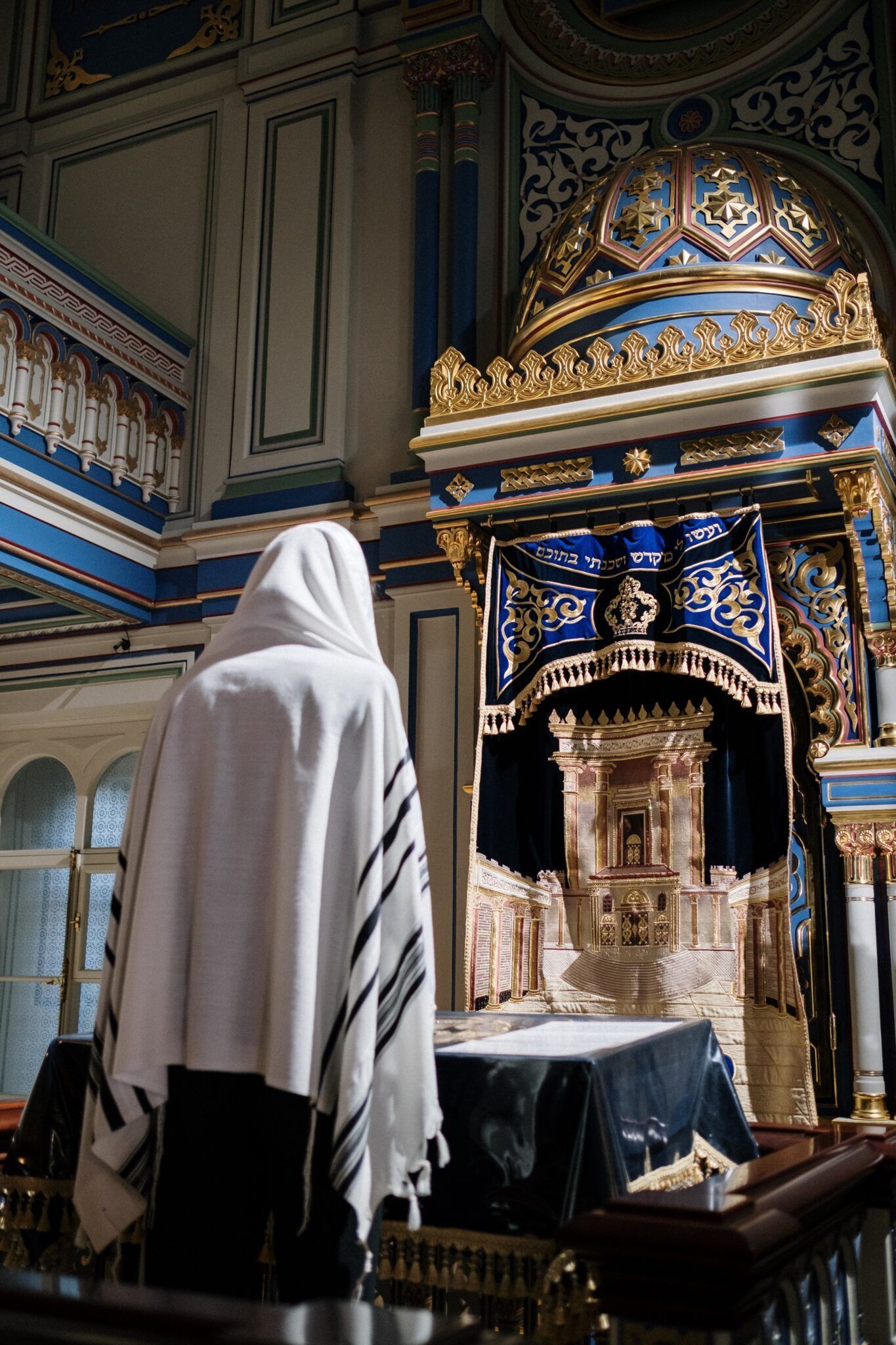Person wearing a white prayer shawl in front of an ornate ark inside a synagogue.