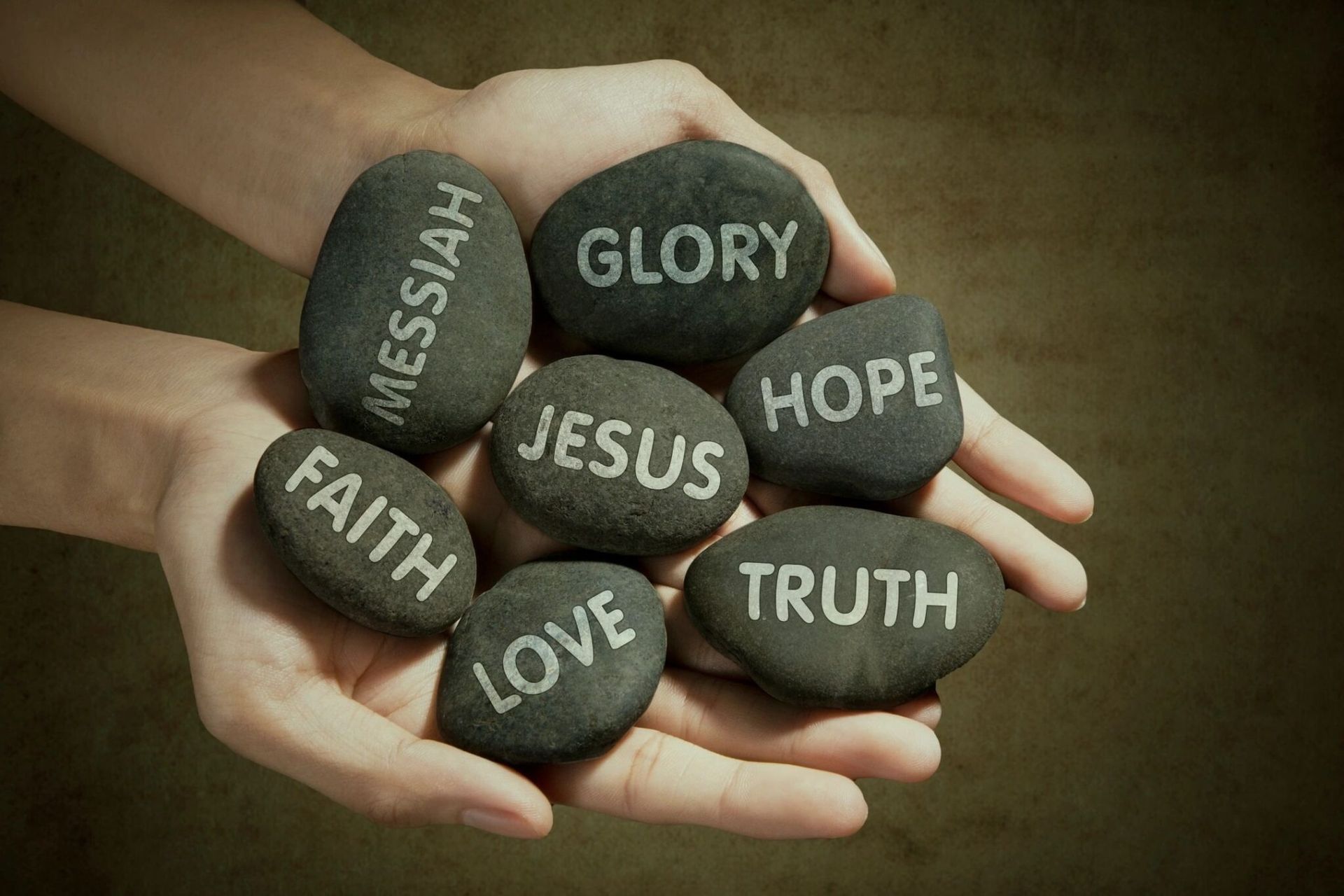 Hands holding seven stones engraved with Christian words: Messiah, Glory, Jesus, Hope, Truth, Love, Faith.