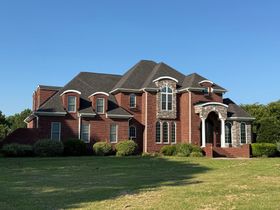 Large red brick suburban house with a dark roof and manicured lawn under a clear blue sky