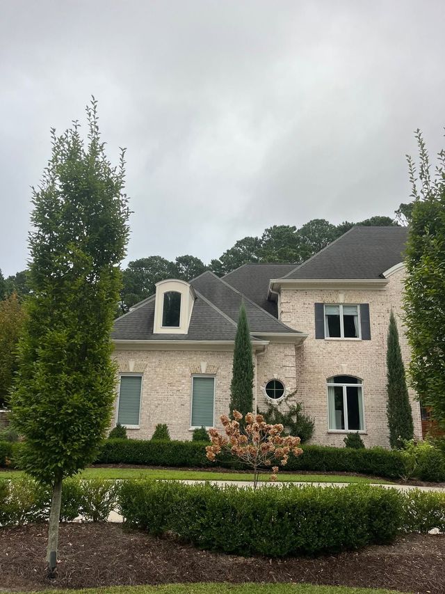 Large stone house with gray roof, manicured hedges, and flowering shrubs under an overcast sky