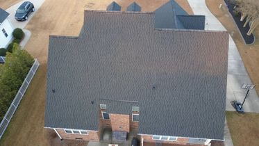 Aerial view of a large gray-shingled suburban house with a driveway and parked car nearby.