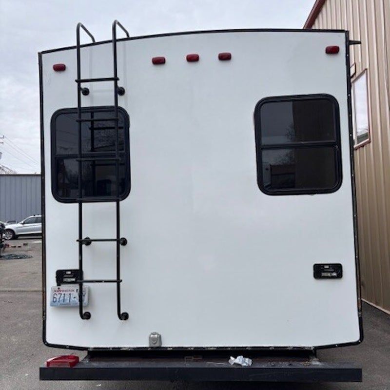 Rear view of a white RV with black trim, windows, and a ladder parked outdoors.