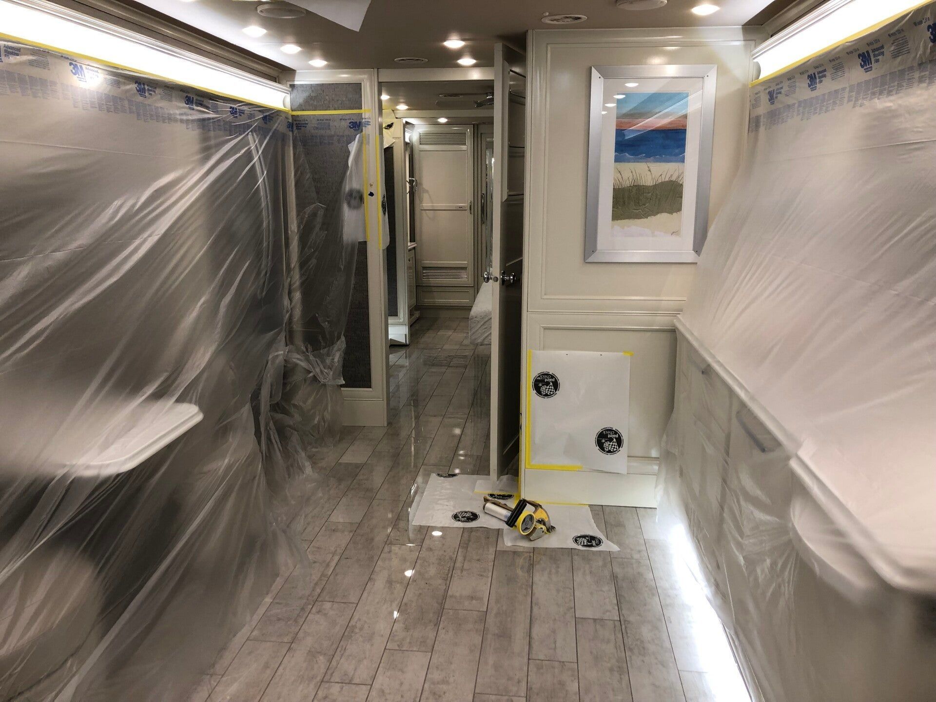 Renovated RV interior wrapped in plastic, with unfinished walls, floor tools, and bright ceiling lights
