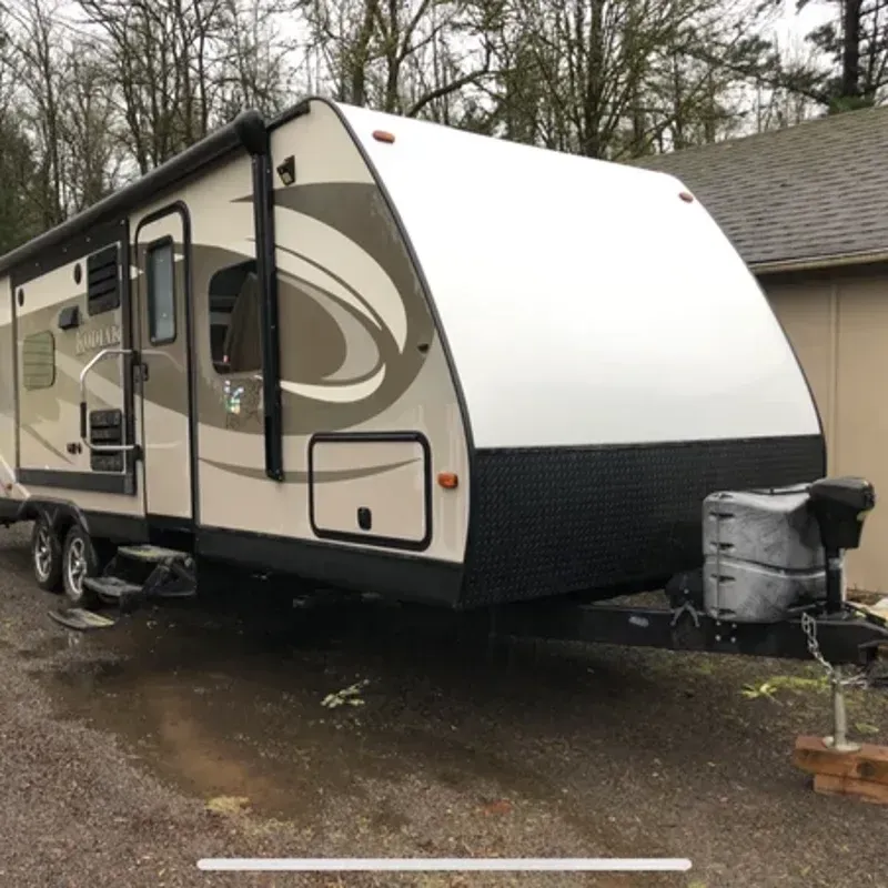 Beige and white travel trailer parked on a wet driveway beside a house.