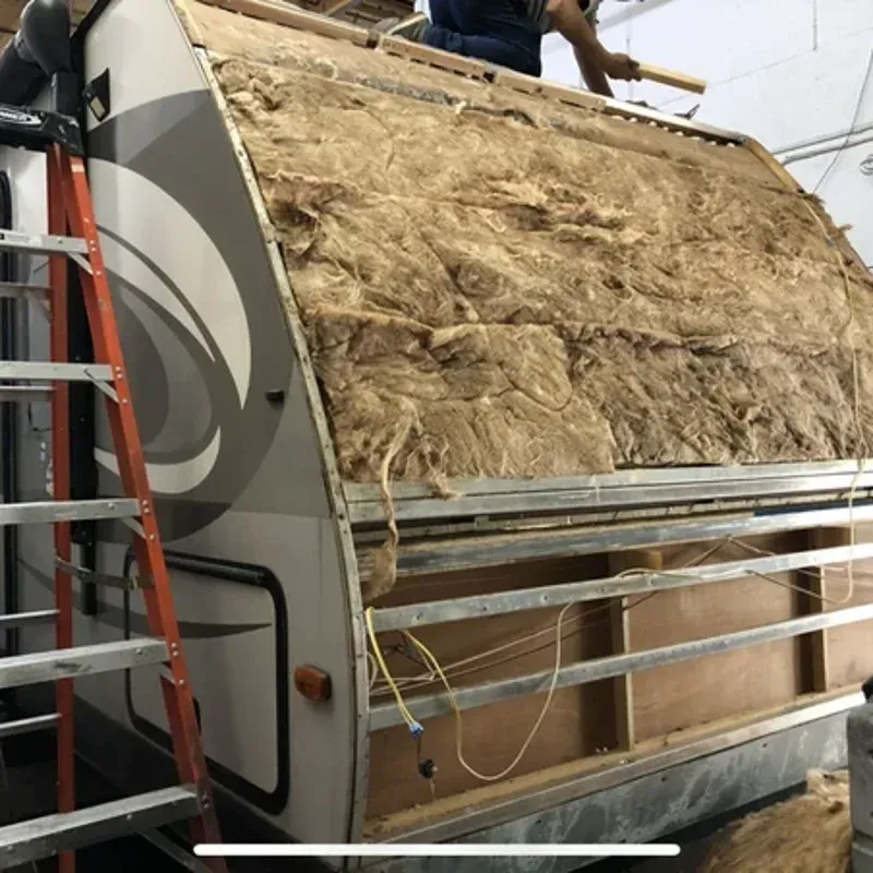 Large insulated trailer or truck panel under construction, with tan insulation exposed and a ladder beside it.