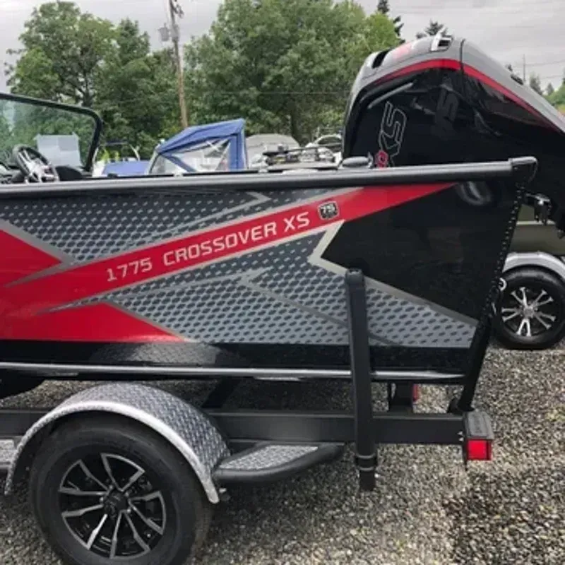 Red and black 1775 Crossover XS fishing boat on a trailer in a gravel lot, with an outboard motor.