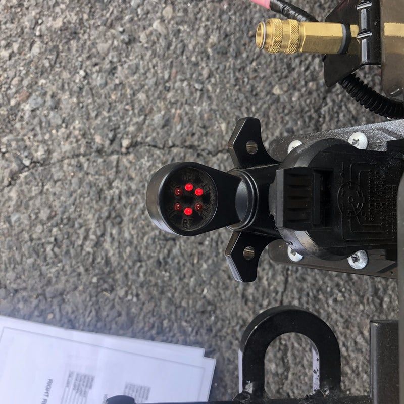 Close-up of a black device with a glowing red LED, resting on pavement near papers and a brass fitting.