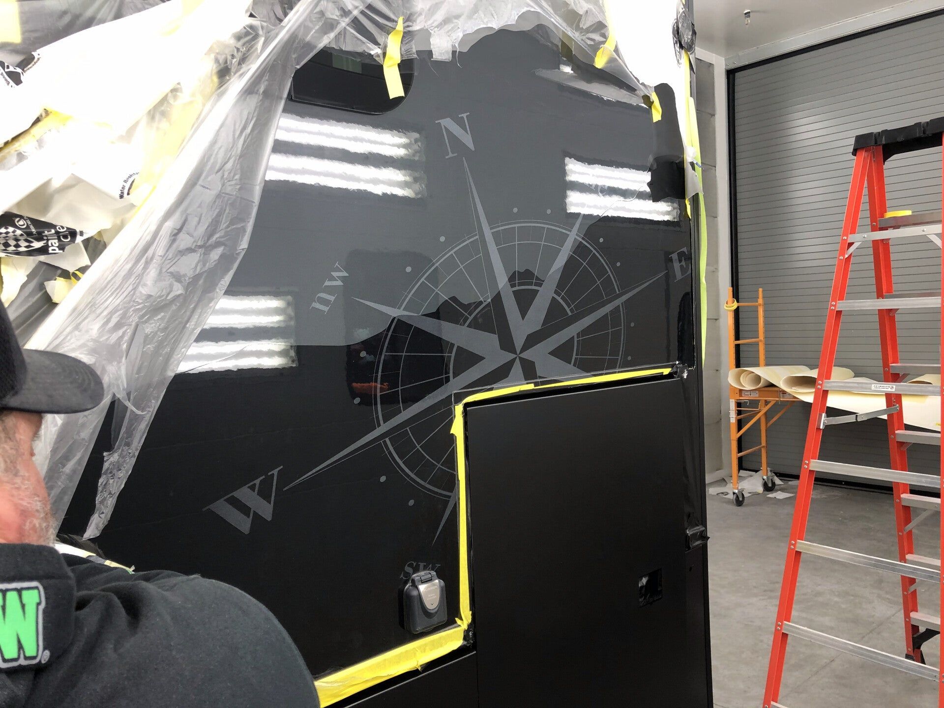A large black boat hull being worked on indoors, with ladder and protective plastic sheeting nearby.