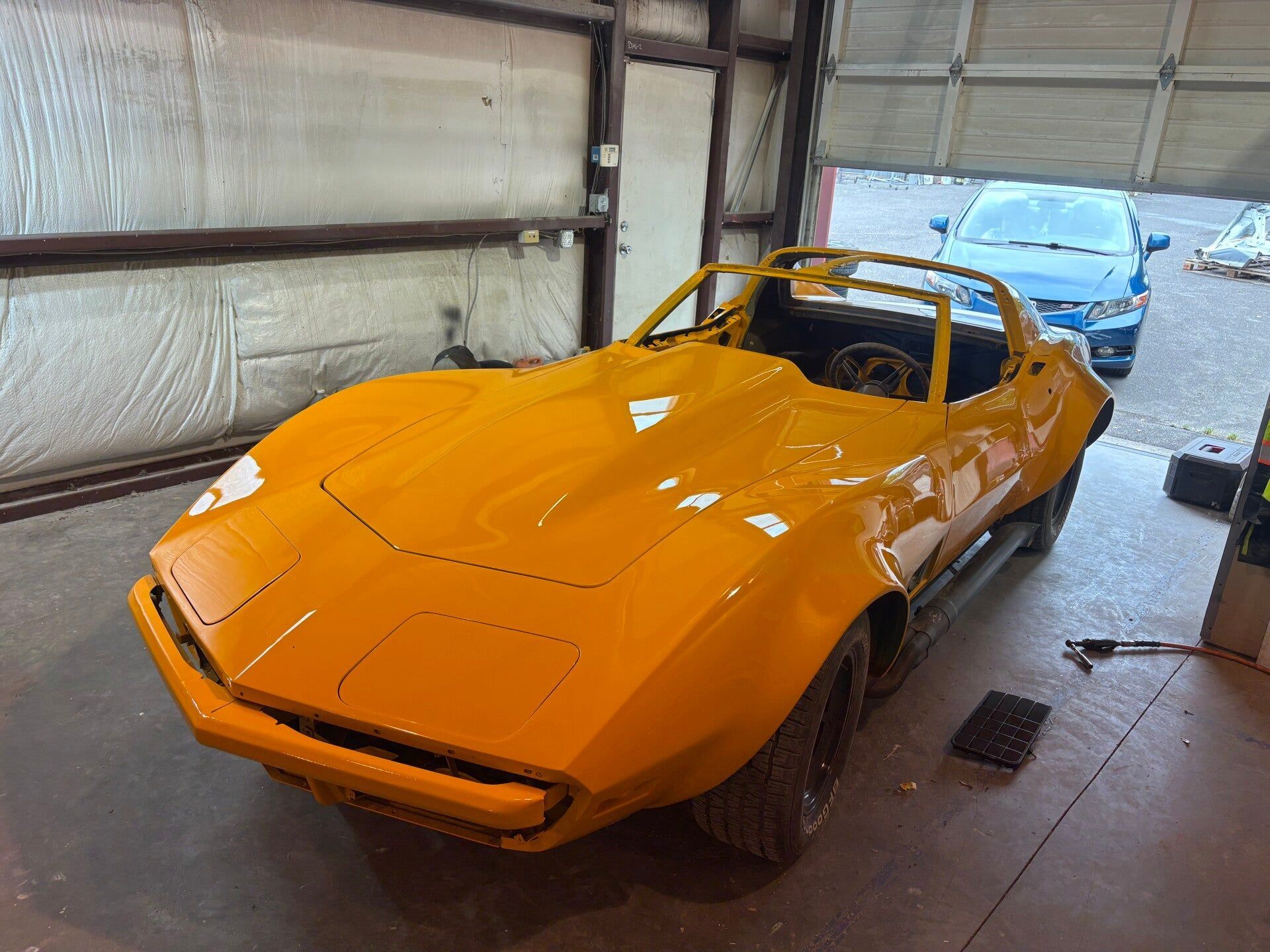 Orange vintage sports car in a garage, shown front-left with a blue car behind it.