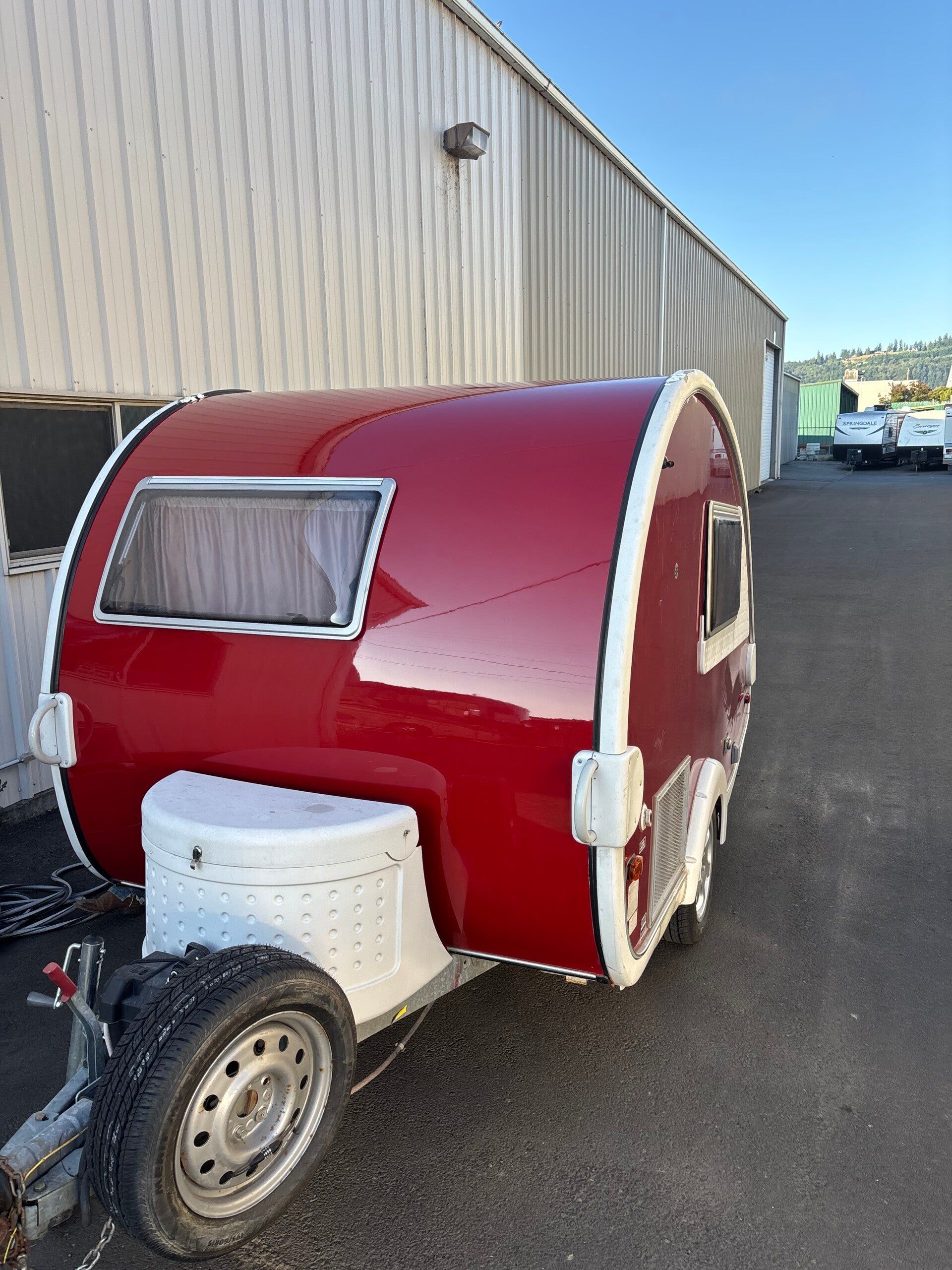 Small red teardrop camper trailer parked beside a warehouse on a paved lot