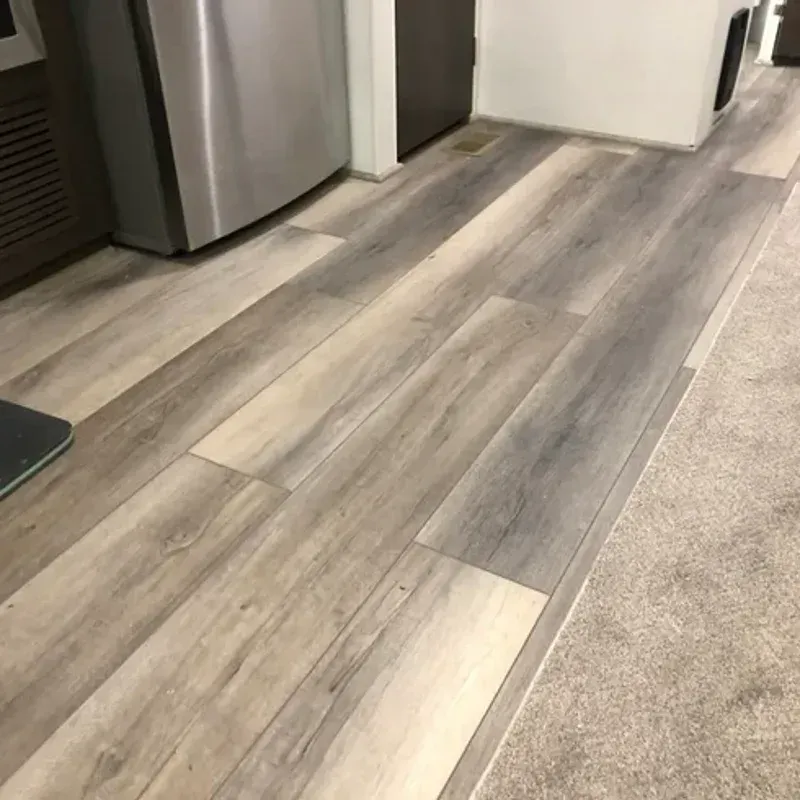 Modern kitchen floor with gray wood-look planks and beige carpet edging, beside stainless steel appliances