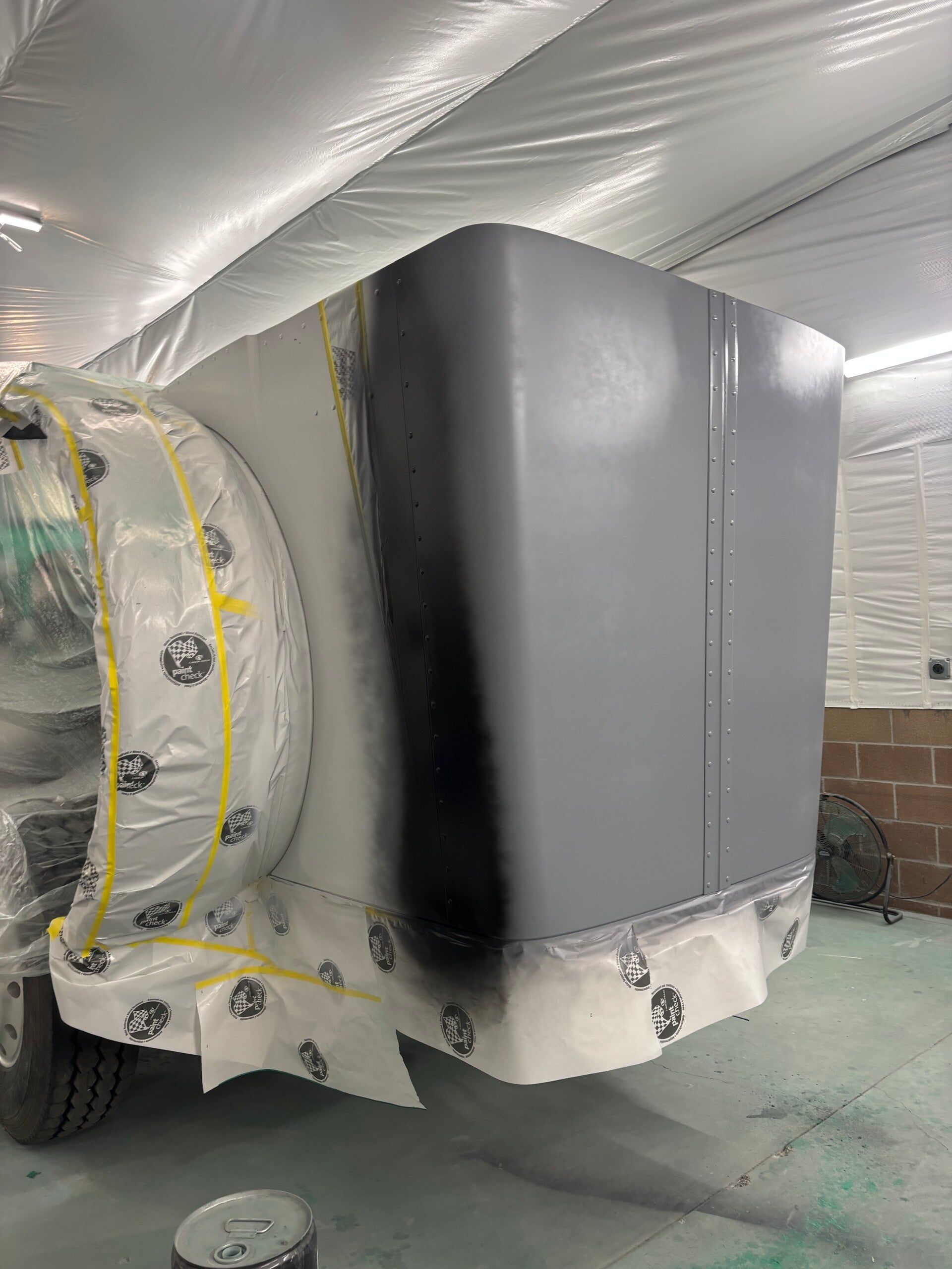 Covered vehicle under masking plastic and tape in a workshop, with a gray primed panel on the rear.