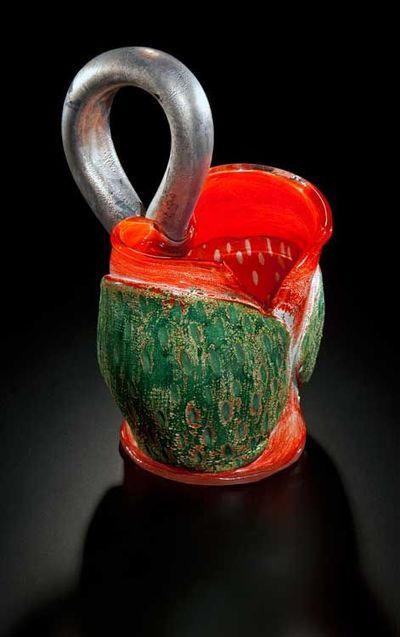 Sculpted art glass. Red and green vessel with silver handle. Black background, reflective surface.