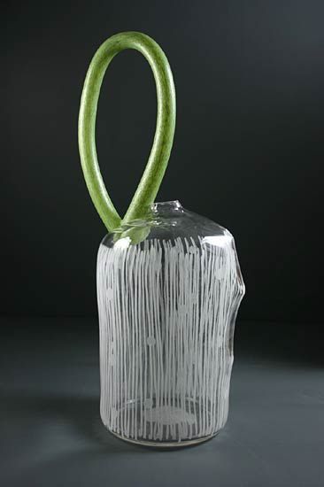 Glass sculpture: clear cylindrical base with vertical white lines, green looping handle. Dark background.
