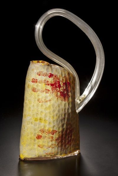 Glass sculpture of a cylindrical vessel with a curved handle. Yellow textured surface with red and yellow splatters.