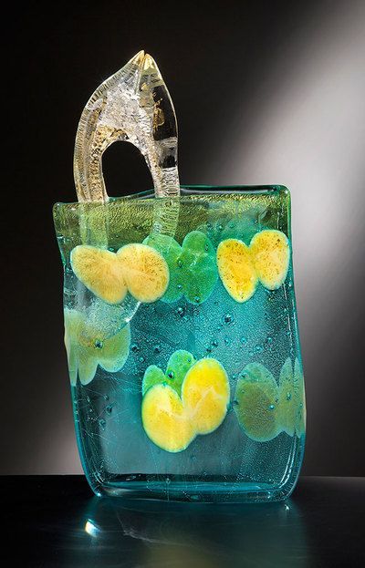 Glass art piece: turquoise vase with yellow and green flower-like shapes, a clear arched handle, on a dark background.
