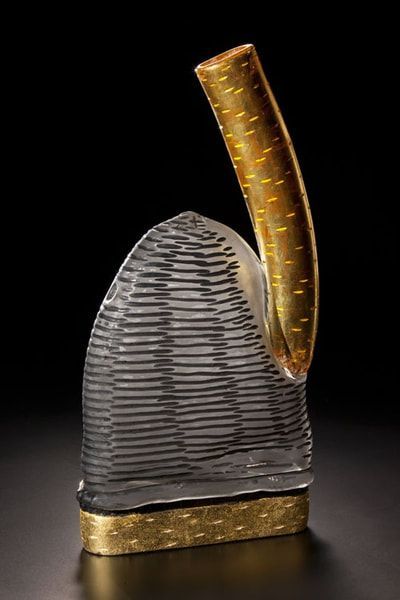 Glass sculpture with a gold, curved top and a textured gray base on a gold rectangular stand.